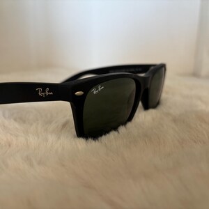 K&ouml;nnte beinhalten: Schwarze Sonnenbrille mit dem Ray-Ban-Logo auf dem B&uuml;gel und der Linse. Die Sonnenbrille hat einen klassischen rechteckigen Rahmen und dunkelgr&uuml;ne Gl&auml;ser. Die Sonnenbrille liegt auf einer wei&szlig;en, flauschigen Oberfl&auml;che.