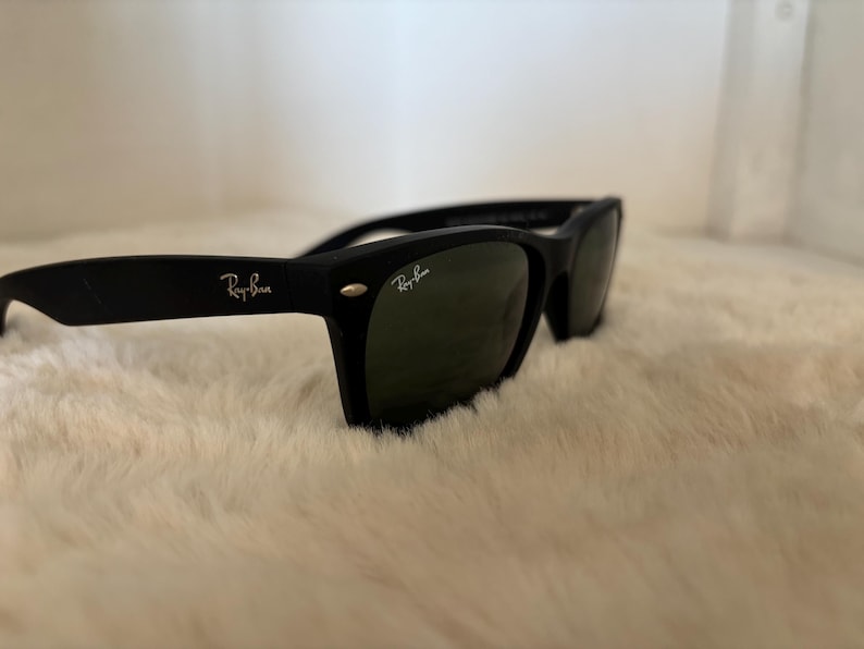 K&ouml;nnte beinhalten: Schwarze Sonnenbrille mit dem Ray-Ban-Logo auf dem B&uuml;gel und der Linse. Die Sonnenbrille hat einen klassischen rechteckigen Rahmen und dunkelgr&uuml;ne Gl&auml;ser. Die Sonnenbrille liegt auf einer wei&szlig;en, flauschigen Oberfl&auml;che.