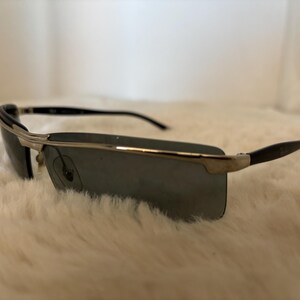May include: Rectangular sunglasses with a silver frame and dark lenses. The temples are black and curve slightly. The sunglasses are set against a white, fluffy background.