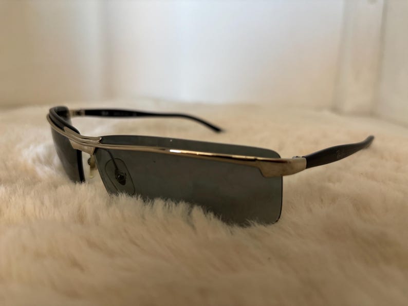 May include: Rectangular sunglasses with a silver frame and dark lenses. The temples are black and curve slightly. The sunglasses are set against a white, fluffy background.