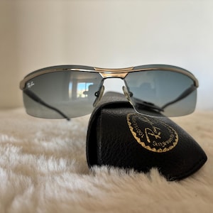 May include: Sunglasses with a gold frame and blue-tinted lenses rest on a black leather case. The sunglasses have a sleek, modern design. The case features a gold emblem.
