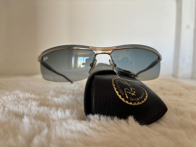 May include: Sunglasses with a gold frame and blue-tinted lenses rest on a black leather case. The sunglasses have a sleek, modern design. The case features a gold emblem.