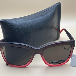 Op de afbeelding: Zonnebril met een donkerbruin montuur dat aan de onderkant overgaat in rood, en een zwarte etui. De glazen zijn donkerbruin van kleur. Een wit label met het woord "JANE" is aan de zonnebril bevestigd.