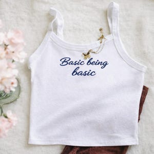 May include: A white vest top with thin straps, embroidered with the navy blue phrase "Basic being basic". A gold-coloured butterfly pendant necklace is draped on the top. Brown ribbed trousers are visible.