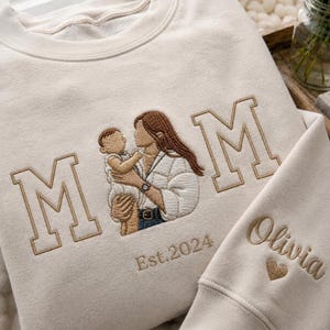 Könnte beinhalten: Cremefarbenes Sweatshirt mit gesticktem Design. Das Design zeigt das Wort „MOM“ in Blockbuchstaben, eine Mutter, die ein Baby hält, und den Text „Est. 2024“. Der Ärmel hat den Namen „Olivia“ und ein farblich passendes gesticktes Herz.