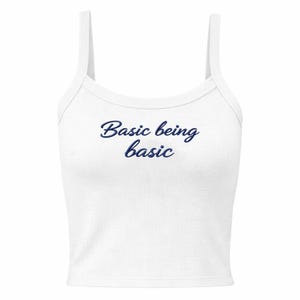 May include: White ribbed vest top with thin straps. The text "Basic being basic" is printed in a dark blue script across the front. The top is a casual style, suitable for layering or warm weather.