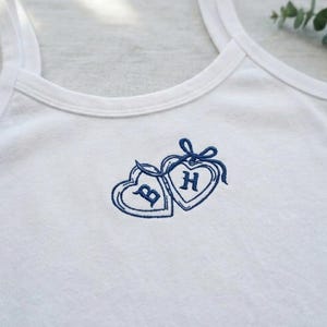 May include: White vest top featuring a blue embroidered design of two linked hearts. Each heart contains a letter, one with a "B" and the other with an "H". A blue ribbon ties the hearts together.