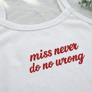 May include: A white vest top featuring red embroidered text that reads "miss never do no wrong" in a cursive script font. The garment has a round neckline and thin shoulder straps, showcasing the detailed stitching of the red lettering.