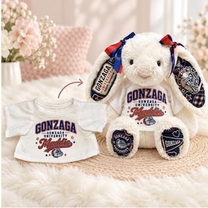 May include: A cream-colored plush bunny with long ears featuring blue and red Gonzaga University patterns, wearing a white t-shirt printed with Gonzaga University and the name Maddie. A matching white t-shirt is placed next to the bunny.