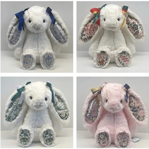 Puede incluir: Cuatro peluches con forma de conejo y orejas largas. Tres conejos son blancos y uno es rosa claro. Cada conejo presenta tela estampada en el interior de las orejas y en los pies, letras griegas bordadas y cintas de colores atadas en las orejas.