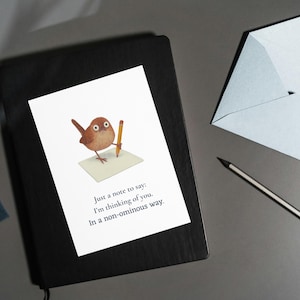 May include: A greeting card with a whimsical illustration of a brown bird holding a pencil, writing on a small pad. The card is on a black notebook, with a light blue envelope and a pencil nearby. Text on the card reads: "Just a note to say: I'm thinking of you. In a non-ominous way."