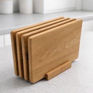 Könnte beinhalten: Ein Satz von vier rechteckigen Holzschneidebrettern in einem natürlichen Holzton, der vertikal in einem passenden Holzhalter ausgestellt wird. Die Bretter haben abgerundete Ecken und sind auf einer hellgrauen Arbeitsplatte angeordnet.