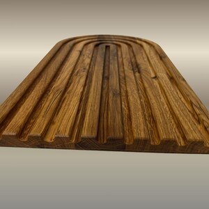 May include: A wooden breadboard with a grooved surface. The board is made of light brown wood and has a rectangular shape with a curved top. The grooves run lengthwise, creating a textured appearance. The board is designed for serving bread.