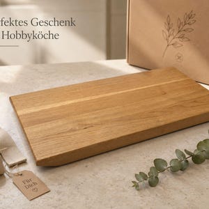 May include: A rectangular light brown wooden cutting board with a smooth finish. Beside it lies a beige ribbon with a small cardboard tag labeled "F&uuml;r Dich" with a heart symbol, a sprig of eucalyptus, and a cardboard box with a leaf illustration.