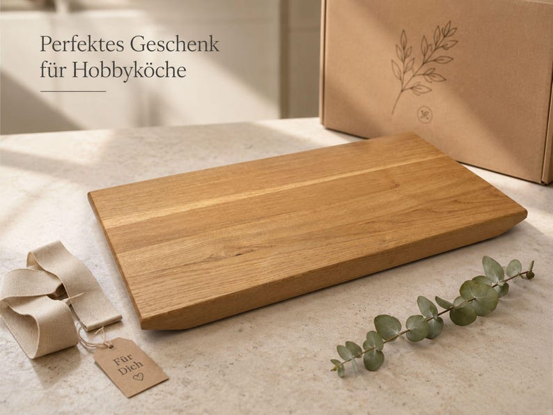 May include: A rectangular light brown wooden cutting board with a smooth finish. Beside it lies a beige ribbon with a small cardboard tag labeled "F&uuml;r Dich" with a heart symbol, a sprig of eucalyptus, and a cardboard box with a leaf illustration.