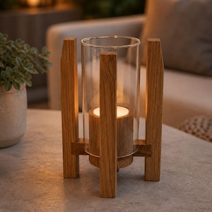 Può includere: Un portacandele in legno con un cilindro di vetro trasparente. Il portacandele ha una base quadrata e quattro supporti verticali in legno. Una candela accesa è all'interno del cilindro di vetro, che proietta un bagliore caldo.