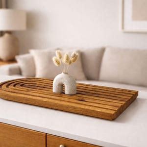 May include: A wooden decorative tray with a carved, arched design, holding a small, off-white vase with dried flowers. The tray is rectangular and has a warm, brown color. The vase has a unique, curved shape.