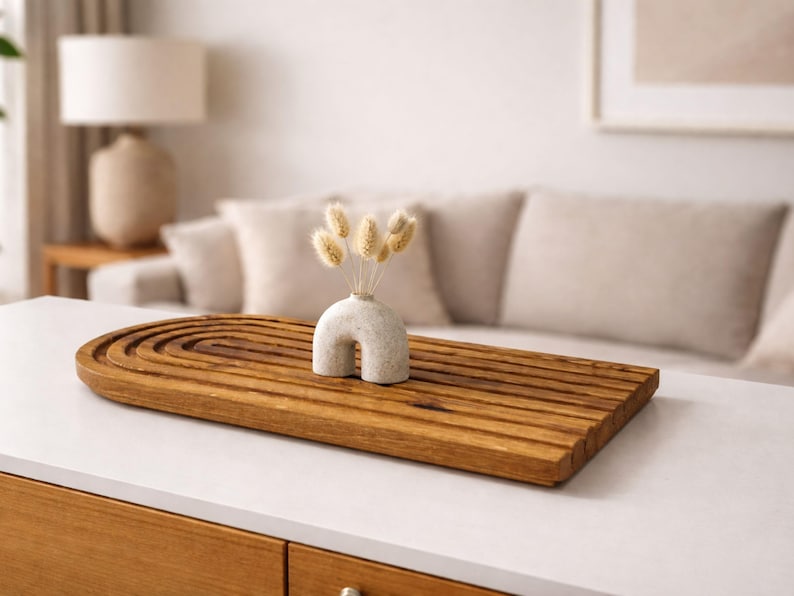 May include: A wooden decorative tray with a carved, arched design, holding a small, off-white vase with dried flowers. The tray is rectangular and has a warm, brown color. The vase has a unique, curved shape.