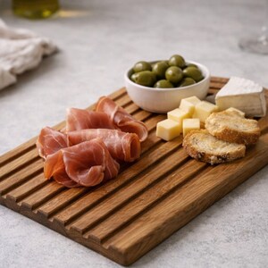 May include: A wooden serving board with grooves, holding prosciutto, olives, cheese cubes, brie, and toasted bread. The board is a warm brown color, perfect for appetizers or a charcuterie spread.