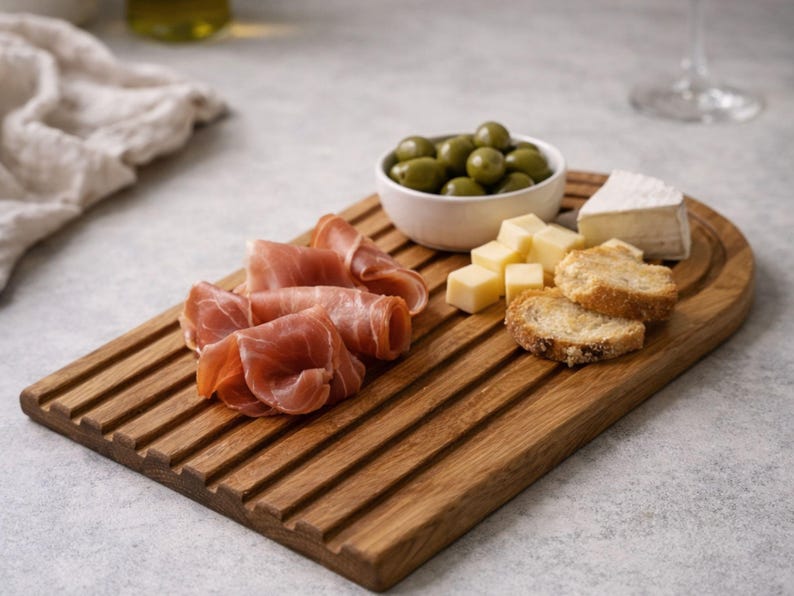 May include: A wooden serving board with grooves, holding prosciutto, olives, cheese cubes, brie, and toasted bread. The board is a warm brown color, perfect for appetizers or a charcuterie spread.