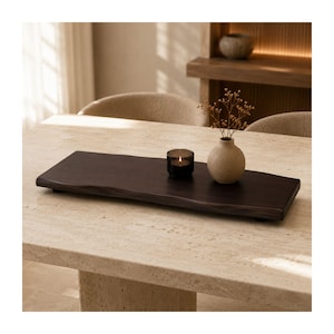 May include: A rectangular dark brown wooden serving tray with a natural live edge, featuring a small round beige ceramic vase containing dried stems and a dark glass candle holder with a lit candle placed on its smooth, flat surface.