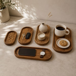 May include: A collection of five oval wooden trays in varying sizes arranged on a surface. The trays hold items including gold rings, a necklace, keys, a key fob, a small white ribbed vase, a candle, a smartphone, a watch, and a mug with a cookie.