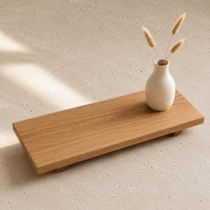 Può includere: Un rialzo rettangolare in legno con finitura a venature naturali, sostenuto da piccoli piedini. Un piccolo vaso in ceramica color bianco sporco contenente tre steli botanici beige, secchi e soffici, poggia sulla superficie in legno.