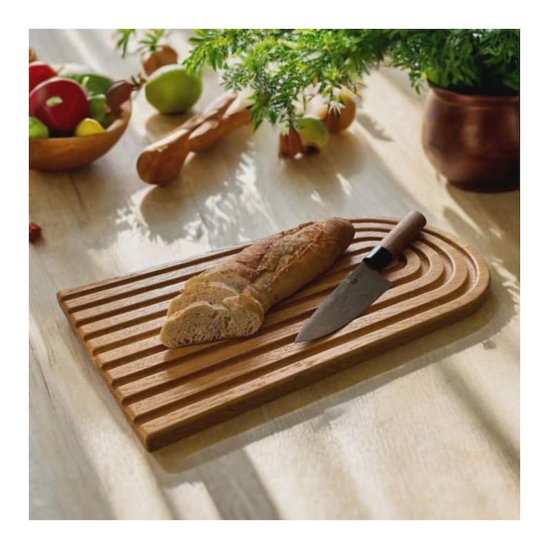 May include: A wooden breadboard with a loaf of sliced bread and a knife. The board has a rectangular shape with parallel grooves and a rounded end. The knife has a wooden handle and a silver blade. The bread is crusty and golden brown.