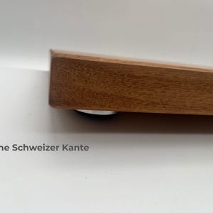 May include: A thick, rectangular wooden board with a natural brown wood grain finish and a straight, blunt edge. A small, circular silver-colored foot is visible underneath the corner. The text "Ohne Schweizer Kante" is printed below the board.