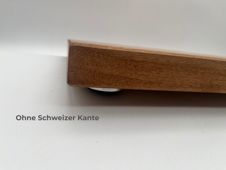 May include: A thick, rectangular wooden board with a natural brown wood grain finish and a straight, blunt edge. A small, circular silver-colored foot is visible underneath the corner. The text "Ohne Schweizer Kante" is printed below the board.