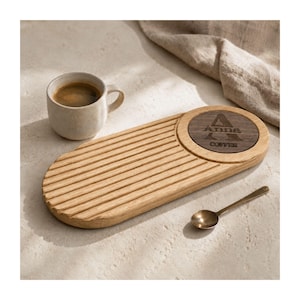 Op de afbeelding: Een houten koffieserveerblad met een gegroefd oppervlak en een cirkelvormige inzet. De inzet toont de naam "Anna" en het woord "Coffee". Een kleine keramische mok met koffie en een gouden lepel liggen naast het dienblad.