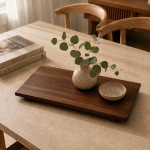 Peut inclure: Un plateau rectangulaire en bois brun foncé doté de quatre petits pieds. Sur le plateau se trouve un vase rond en céramique beige moucheté contenant une branche d'eucalyptus verte, ainsi qu'un petit bol en céramique beige assorti.