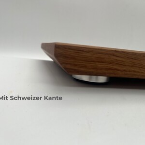 May include: A wooden board with a beveled edge, known as a Swiss edge, featuring a visible wood grain pattern. The board is elevated by a small, circular, silver-colored metal foot with a black base. The text Mit Schweizer Kante is visible.