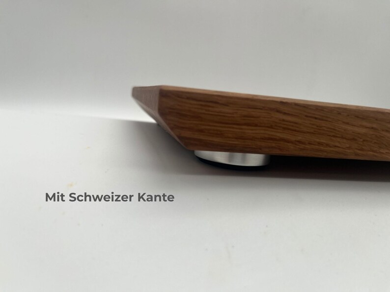 May include: A wooden board with a beveled edge, known as a Swiss edge, featuring a visible wood grain pattern. The board is elevated by a small, circular, silver-colored metal foot with a black base. The text Mit Schweizer Kante is visible.