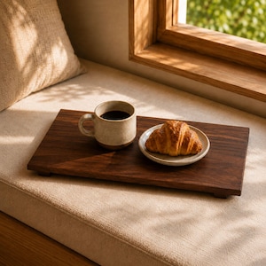 Könnte beinhalten: Ein rechteckiges Serviertablett aus dunkelbraunem Holz mit kurzen Füßen, darauf eine beige Keramiktasse mit schwarzem Kaffee und ein kleiner weißer Teller mit einem goldbraunen, blättrigen Croissant. Das Holz hat eine glatte Maserung.