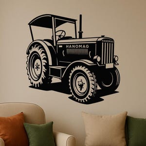 Op de afbeelding: Zwart-wit muurkunst van een vintage Hanomag-tractor. De tractor heeft grote spaakwielen, een cabine en de naam Hanomag. Het kunstwerk is op een neutrale muur gemonteerd, met een stoel en een bank op de voorgrond.