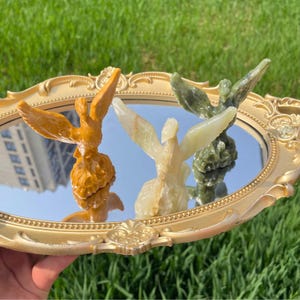 May include: Three carved stone bird figurines in shades of yellow, cream, and green are displayed on a gold-framed mirrored tray. The birds have outstretched wings and are set against a backdrop of green grass.
