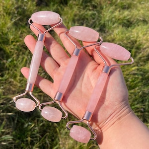 Puede incluir: Cuatro rodillos faciales de cuarzo rosa. Cada rodillo presenta una piedra rosa claro con un mango cilíndrico y dos cabezas redondeadas, conectadas por un marco de metal dorado. Los rodillos se sostienen en una mano sobre un fondo de hierba verde.