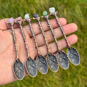 May include: Six decorative spoons with ornate bronze-colored handles and floral designs on the spoon bowls. Each spoon is topped with a different colored gemstone: purple, pink, clear, blue, green, and clear.