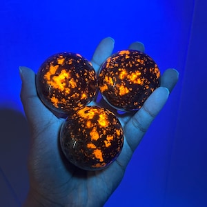 Könnte beinhalten: Drei kugelförmige Objekte mit dunkelbrauner Basis und leuchtend orangefarbenen Sprenkeln werden in einer Hand gehalten. Die Kugeln sind beleuchtet und erzeugen einen Leuchteffekt vor blauem Hintergrund. Die Objekte scheinen dekorativ oder sammelbar zu sein.