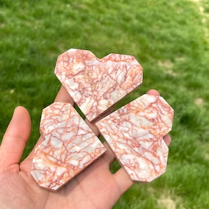 Puede incluir: Tres piedras en forma de corazón con un diseño geométrico. Las piedras son blancas con vetas rojas, sostenidas en una mano sobre un fondo de hierba verde. Los corazones tienen un acabado pulido.