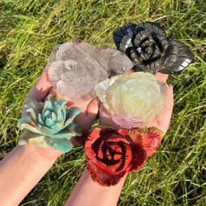 Puede incluir: Una colección de rosas de piedra talladas en varios colores, incluyendo transparente, negro, verde, rojo y amarillo. Las rosas se sostienen en manos abiertas sobre un fondo de hierba verde. Los intrincados detalles de los pétalos son visibles.