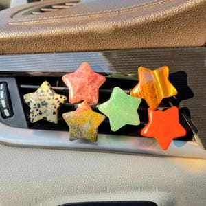 May include: A collection of six star-shaped car air fresheners in various colors and patterns. The stars include speckled, red, green, orange, and yellow-brown designs. They are placed on a car's air vent.
