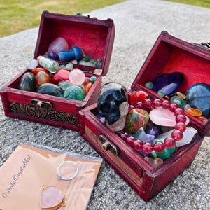 May include: Two open, red wooden treasure chests filled with colorful gemstones and crystals. One chest contains a black crystal skull. A small package with a heart-shaped pendant and the text "OrientalCrystalArt" is in the foreground.