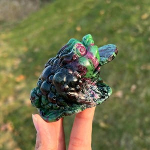May include: A detailed frog sculpture in shades of green, purple, and black. The frog's textured skin and bulging eyes are visible. The sculpture is held in a person's hand against a blurred green background.