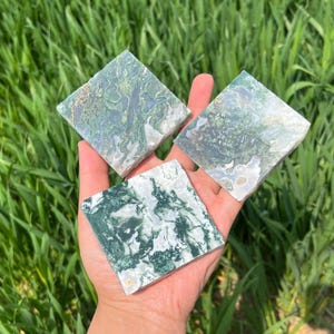 Puede incluir: Tres piedras cuadradas de ágata musgosa con patrones verdes y blancos se sostienen en una mano. Las piedras tienen un aspecto natural, con variaciones de color y textura. El fondo es hierba verde borrosa.