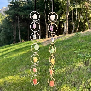 May include: Two hanging crystal wind chimes. Each chime features a series of silver-colored rings, each holding a different colored raw crystal. The crystals include white, purple, green, yellow, and red-orange stones. The chimes hang against a backdrop of green grass and trees.