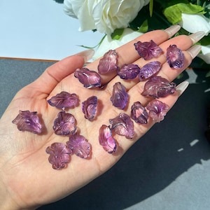 May include: A collection of small, carved amethyst gemstone flower shapes. The translucent purple stones are intricately detailed, with a range of shades from light lavender to deep violet. The gemstones are displayed on a light background.