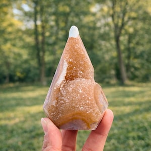 Può includere: Una mano che tiene un cristallo di geode con una parte superiore a punta. Il cristallo è un mix di colori arancione, marrone e bianco. La geode ha una superficie ruvida e strutturata e un aspetto liscio e lucido in alcune aree. Lo sfondo è una vista sfocata di alberi ed erba verdi.