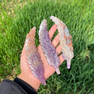 Peut inclure: Trois plumes en pierre sculptée aux tons violets, orange et blancs sont présentées. Les plumes sont tenues dans une main sur fond d'herbe verte. Les plumes présentent des veinures détaillées.